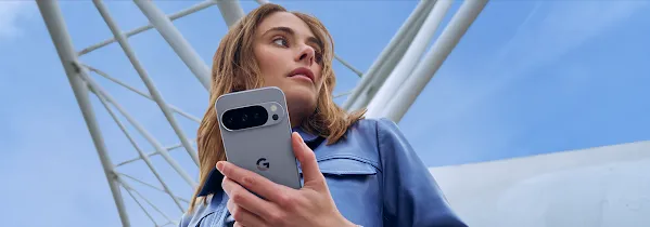 Da una prospettiva dal basso, una persona con una giacca blu tiene in mano uno smartphone Pixel grigio. Sullo sfondo si vedono un cielo azzurro brillante e travi architettoniche bianche.